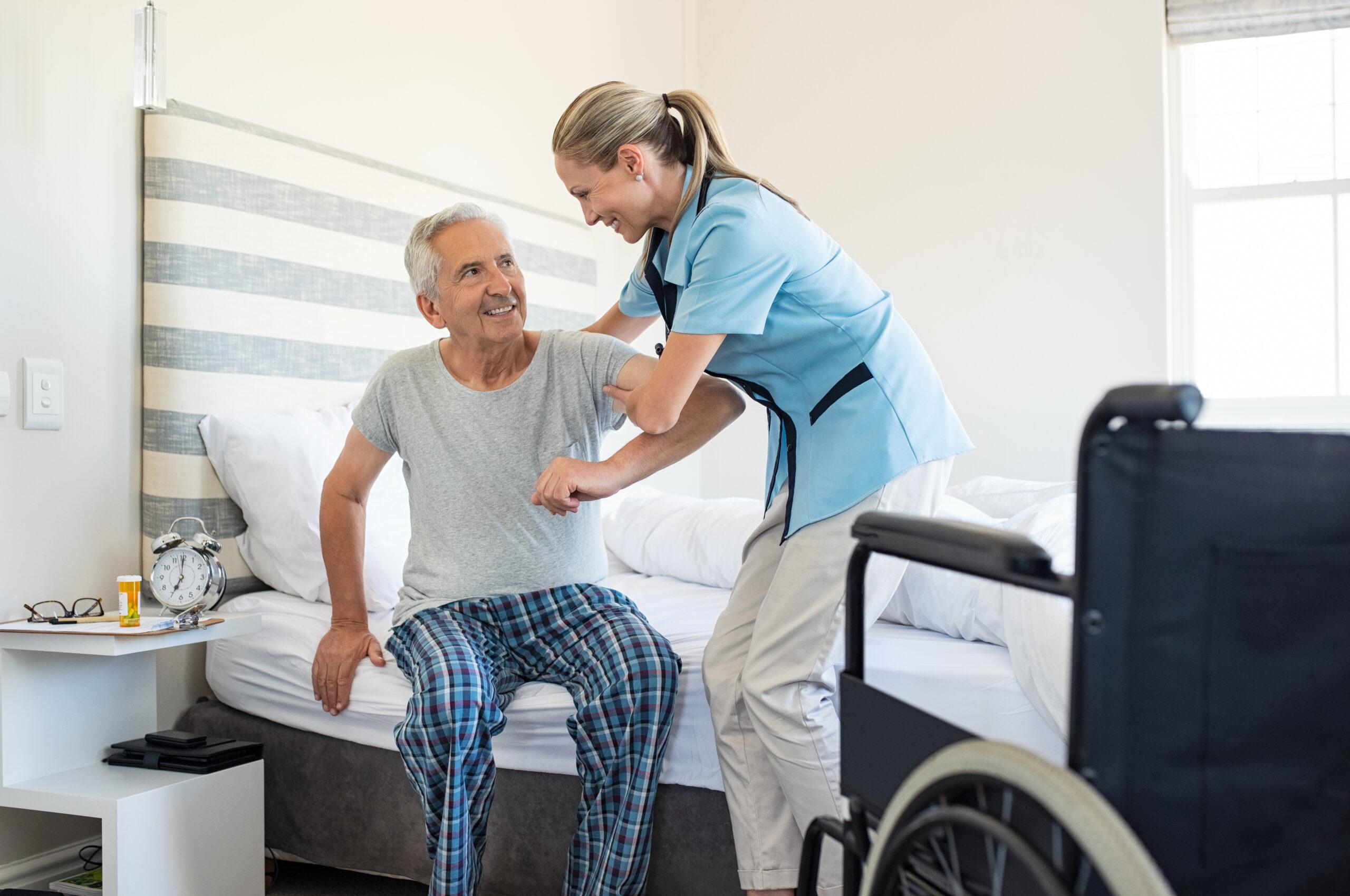 Smiling nurse assisting senior man to get up from bed. Caring nurse supporting patient while getting up from bed and move towards wheelchair at home. Helping elderly disabled man standing up in his bedroom. — Photo